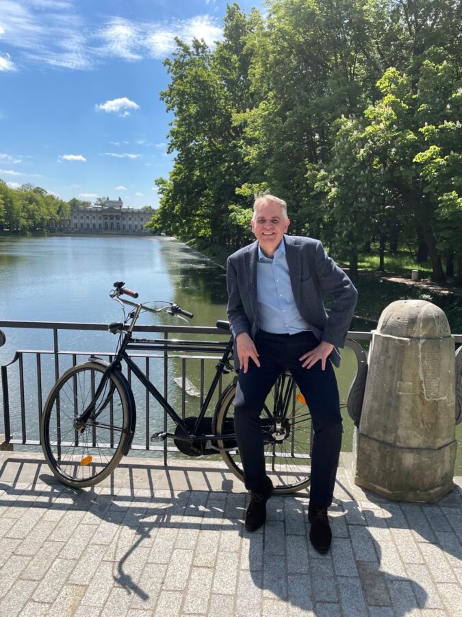 Ambasador Królestwa Niderlandów w Polsce Jennes de Mol pozuje z rowerem na tle rzeki i pałacu.